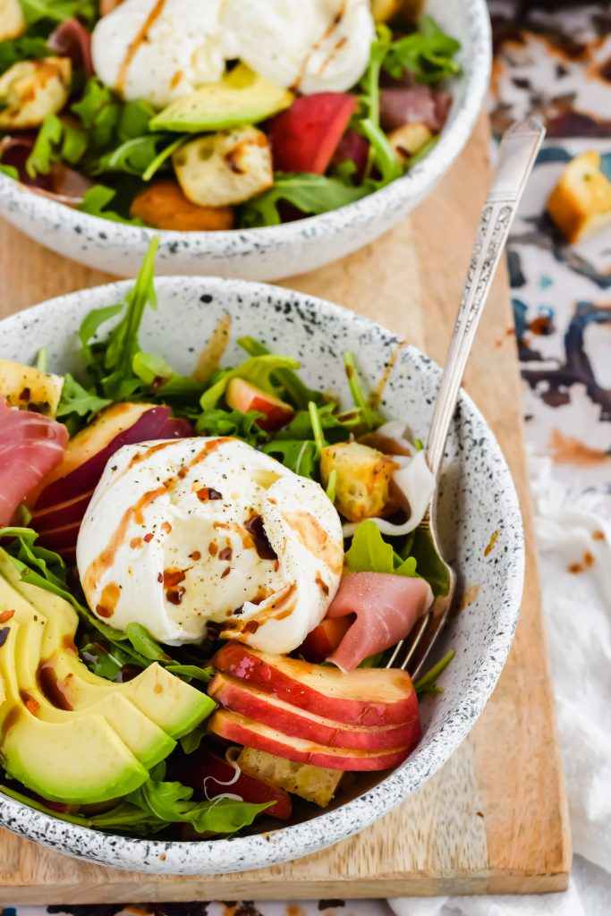 close up of white ceramic salad bowl filled with arugula, peaches, avocado slices, and burrata cheese with balsamic dressing
