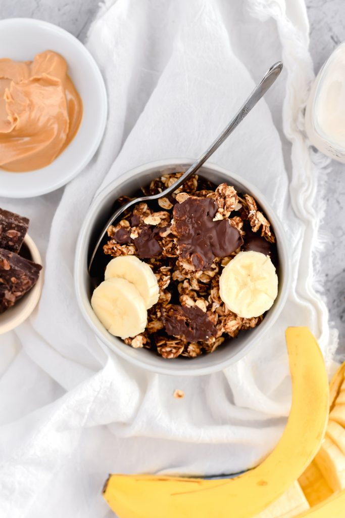 small white bowl of peanut butter chocolate granola on white linen topped with three banana slices  with banana, bowl of peanut butter, and bowl of chocolate in background 