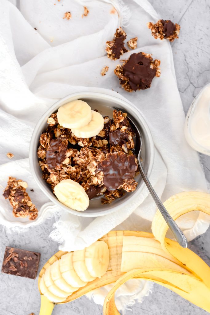 small white bowl of peanut butter chocolate granola on white linen topped with three banana slices along side beside a sliced banana in its peel