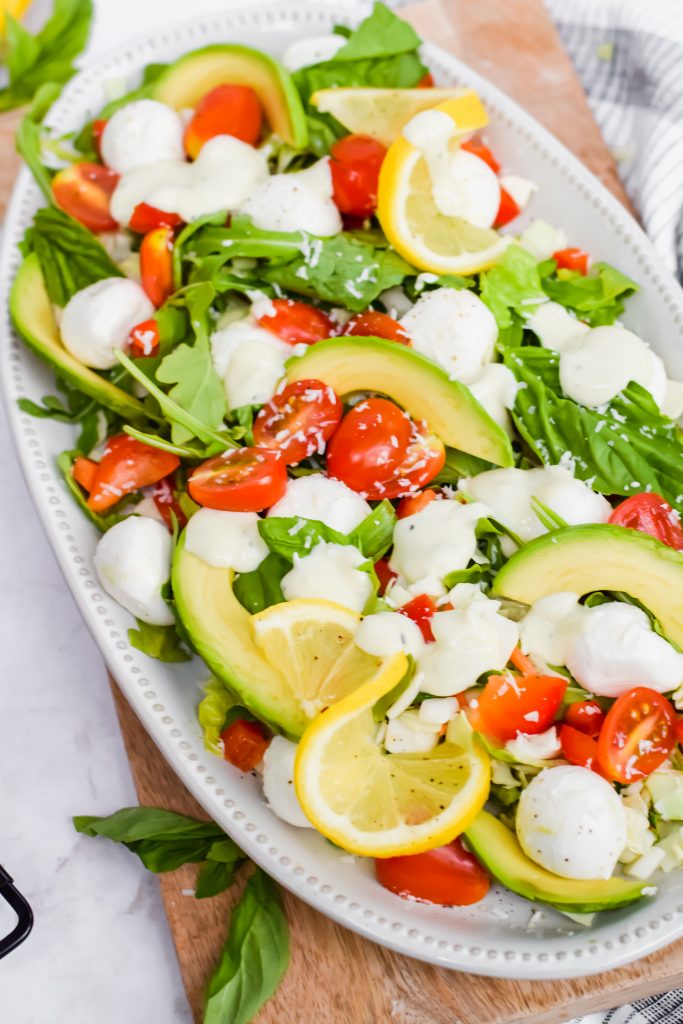 close up angled shot of lemon basil caprese salad on white serving tray on top of wood cutting board with white background with additional salad ingredients scattered around