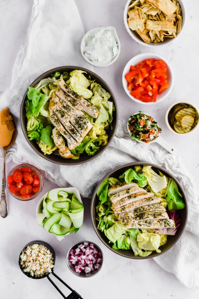 two bowls of salad topped with chicken on white background alongside additional smaller bowls of mediterranean salad ingredientts including cucumber, tomatoes, pickles, onion, feta, peppers, and pita chips