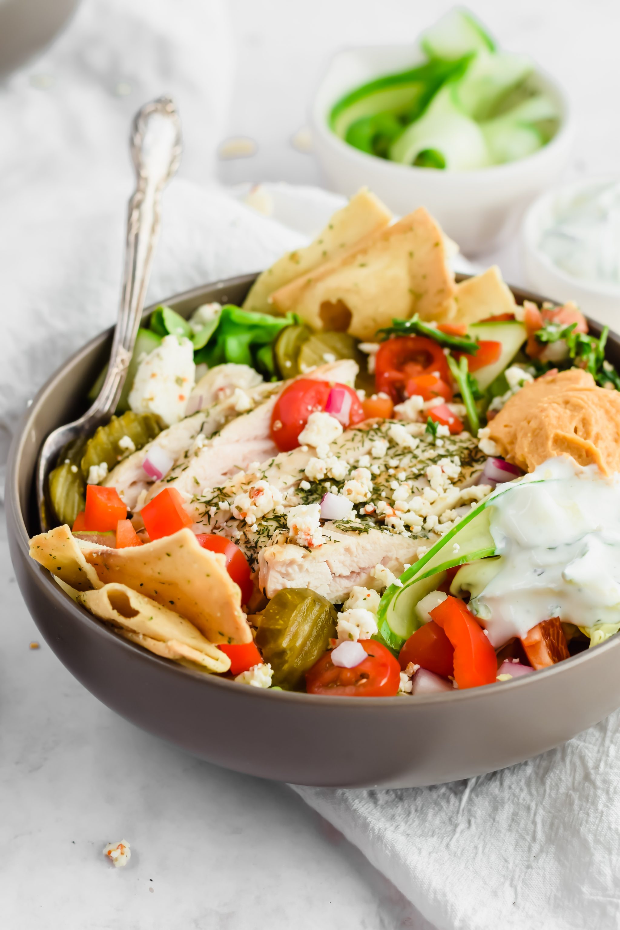 45 degree close up image of loaded salad bowl with sliced grilled chicken, pita chips, tomatoes, red onions, pickles, dollop of hummus, tabbouleh, and tzatziki dressing