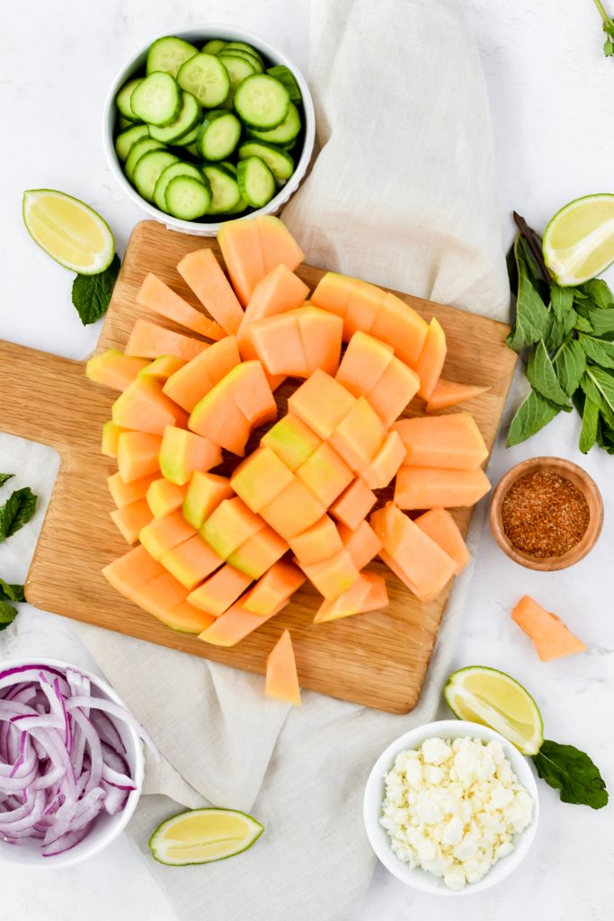 sliced cantaloupe on wood cutting board and other ingredients for the salad surrounding it