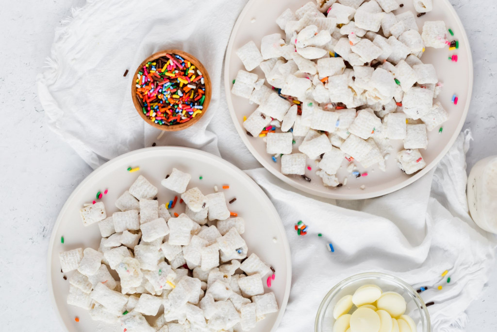 two plates of funfetti puppy chow plated on white dishes and sprinkles surrounding them 