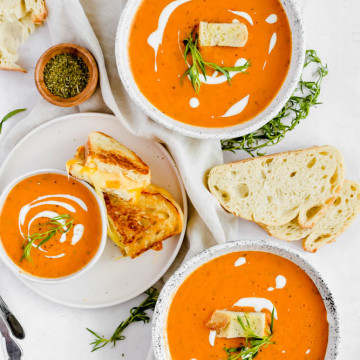 three bowls of tomato soup with grilled cheeses and slices of bread on white background