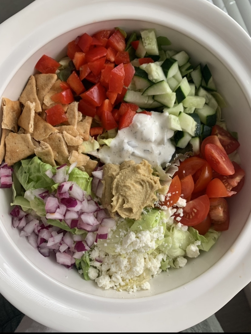 bowl of salad topped with red pepper, tomatoes, cucumber, pita chips, red onion, and hummus