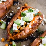 mediterranean sweet potato split open and topped with tzatiki, cucumbers, fresh dill and chickpeas on parchment.