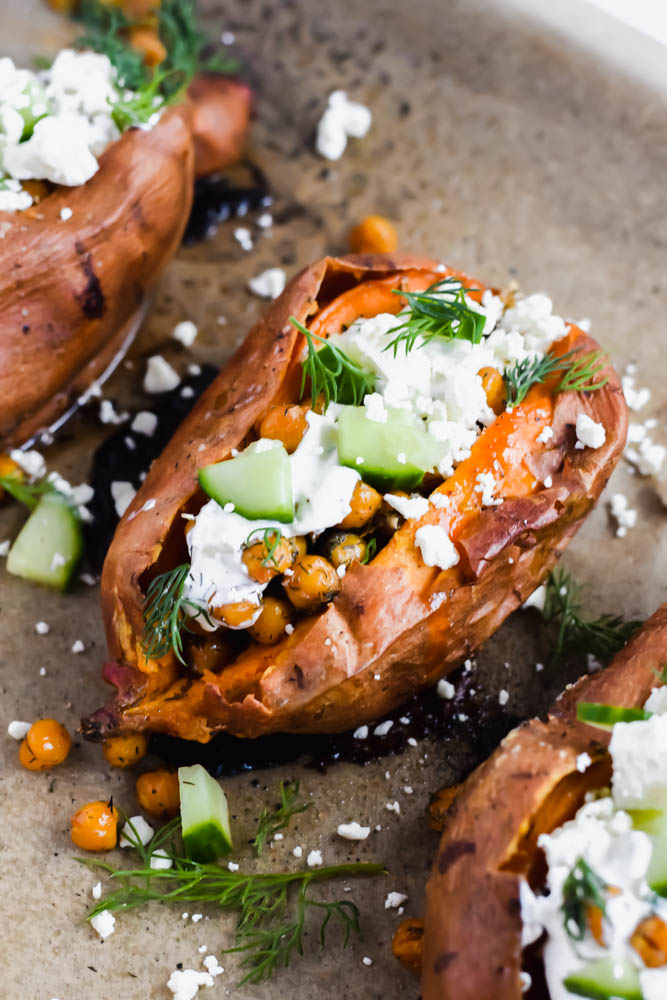 baked sweet potato split open and topped with tzatiki sauce, diced cucumbers, fresh dill and roasted chickpeas on parchment paper