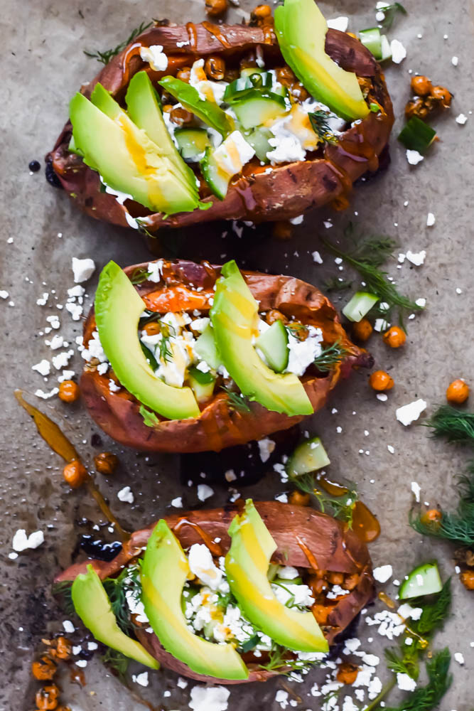 three baked sweet potato loaded and garnished with fresh avocado and spicy honey on gray backdrop surrounded by additional toppings