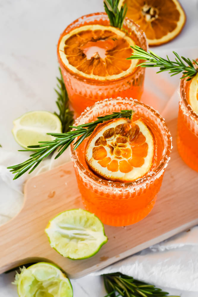 three Italian Margaritas garnished with fresh rosemary and dried oranges and limes on the side