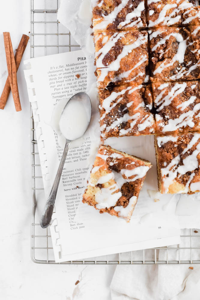 cinnamon coffee cake cut in squares with spoon filled with icing on ripped out sheets of a cookbook and garnished with cinnamon sticks and icing