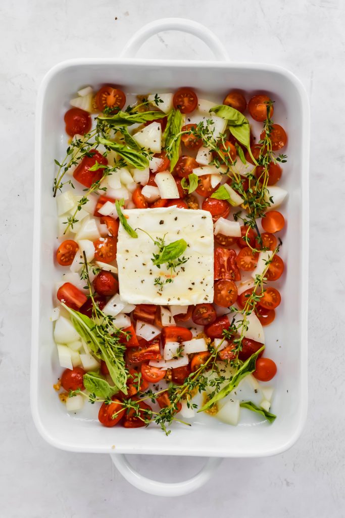Large white baking dish filled with raw onion, sliced cherry tomatoes, thyme sprigs, basil leaves and a block of feta in the center on white backdrop