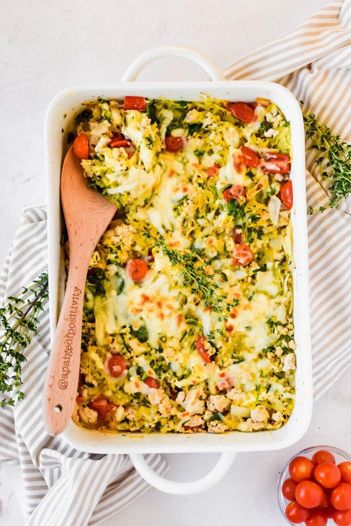 Large white baking dish with baked feta spaghetti squash with wooden spoon in dish on striped linen backdrop with fresh thyme sprig and small bowl of cherry tomatoes along side of baking dish