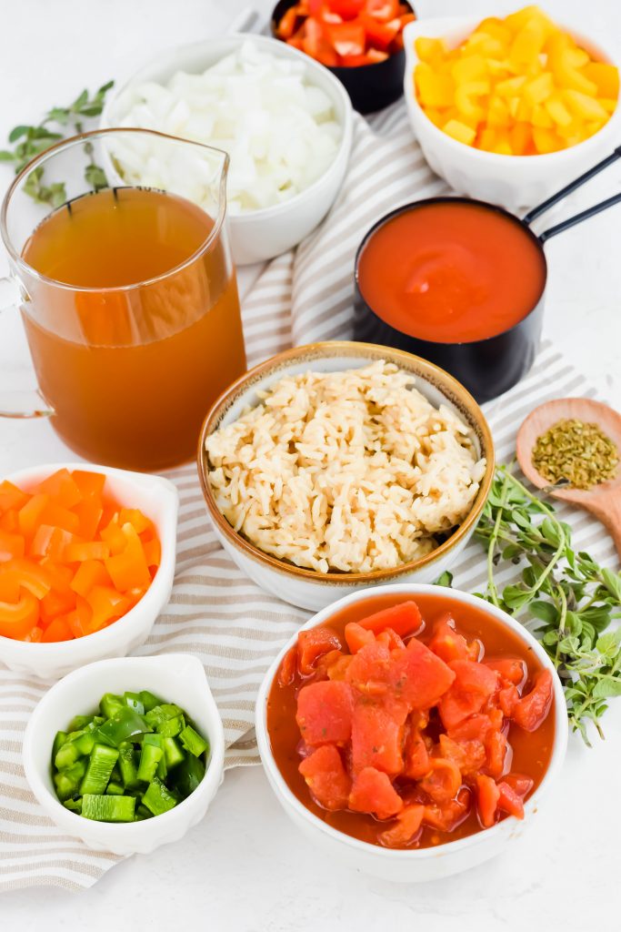 Multiple bowls of separated ingredients including vegetables, rice, broth, and spices on striped linen backdrop