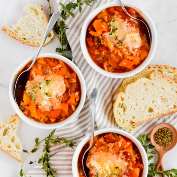 Three white bowls of Vegetarian Stuffed Pepper Soup with silver spoons garnished with cheese and fresh thyme with spoons in the bowls on a striped tan and white linen with sprigs of thyme and fresh bread and thyme sprigs surrounding
