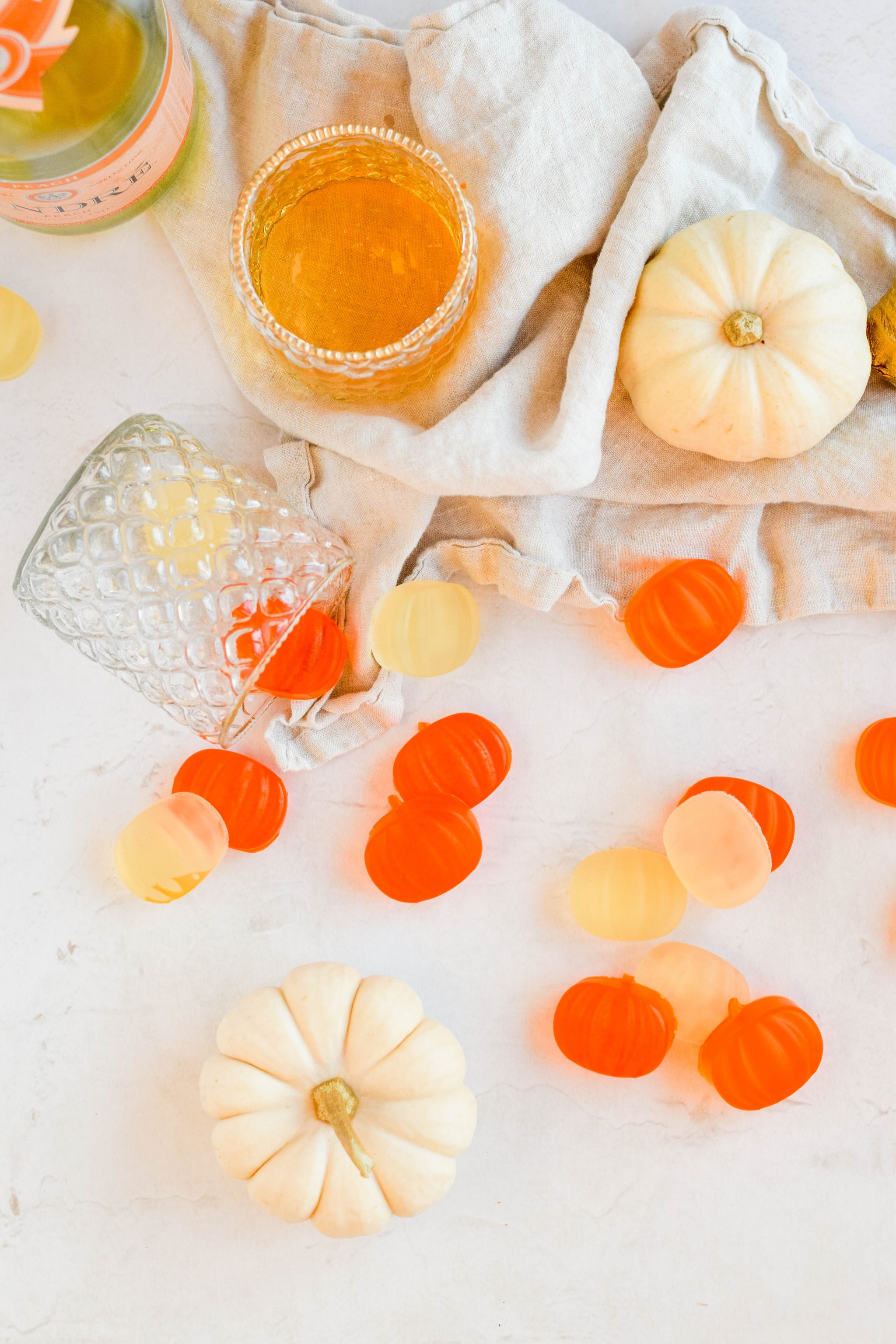 pumpkin shaped champagne gummies spread out with cream pumpkins and glass of champagne on white background