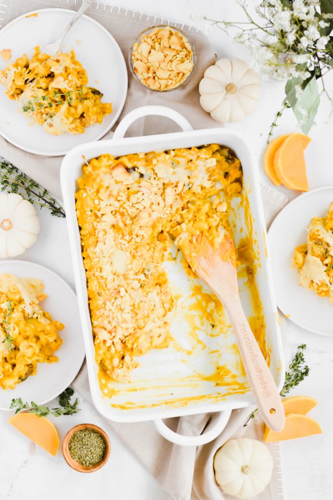 Large white dish half filled with roasted butternut squash mac n' cheese on neutral backdrop surrounded by herb springs, miniature pumpkins, dish ingredients, and plates filled with mac n' cheese