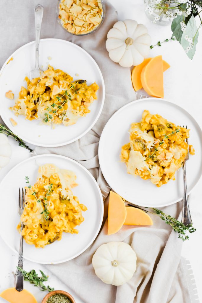 Three servings of Roasted Butternut Squash Mac 'n Cheese dished onto white serving plates on neutral back drop surrounded by white pumpkin, herb sprigs, and dish ingredients