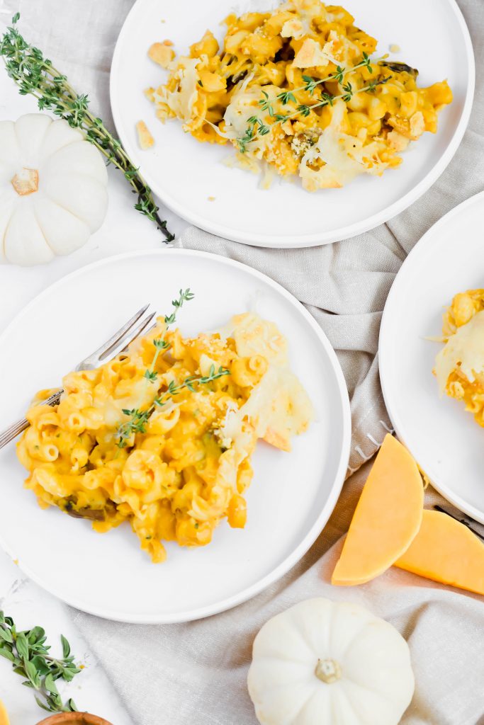 Close up of two servings of Butternut Squash Mac 'n Cheese on white serving plates topped with fresh thyme sprigs on neutral background with forks and surrounded by miniature pumpkin and recipe ingredients