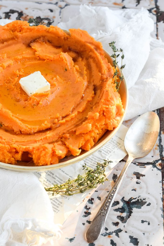 savory mashed sweet potatoes in a white bowl topped with a pat of butter on vintage background beside a silver spoon and fresh thyme