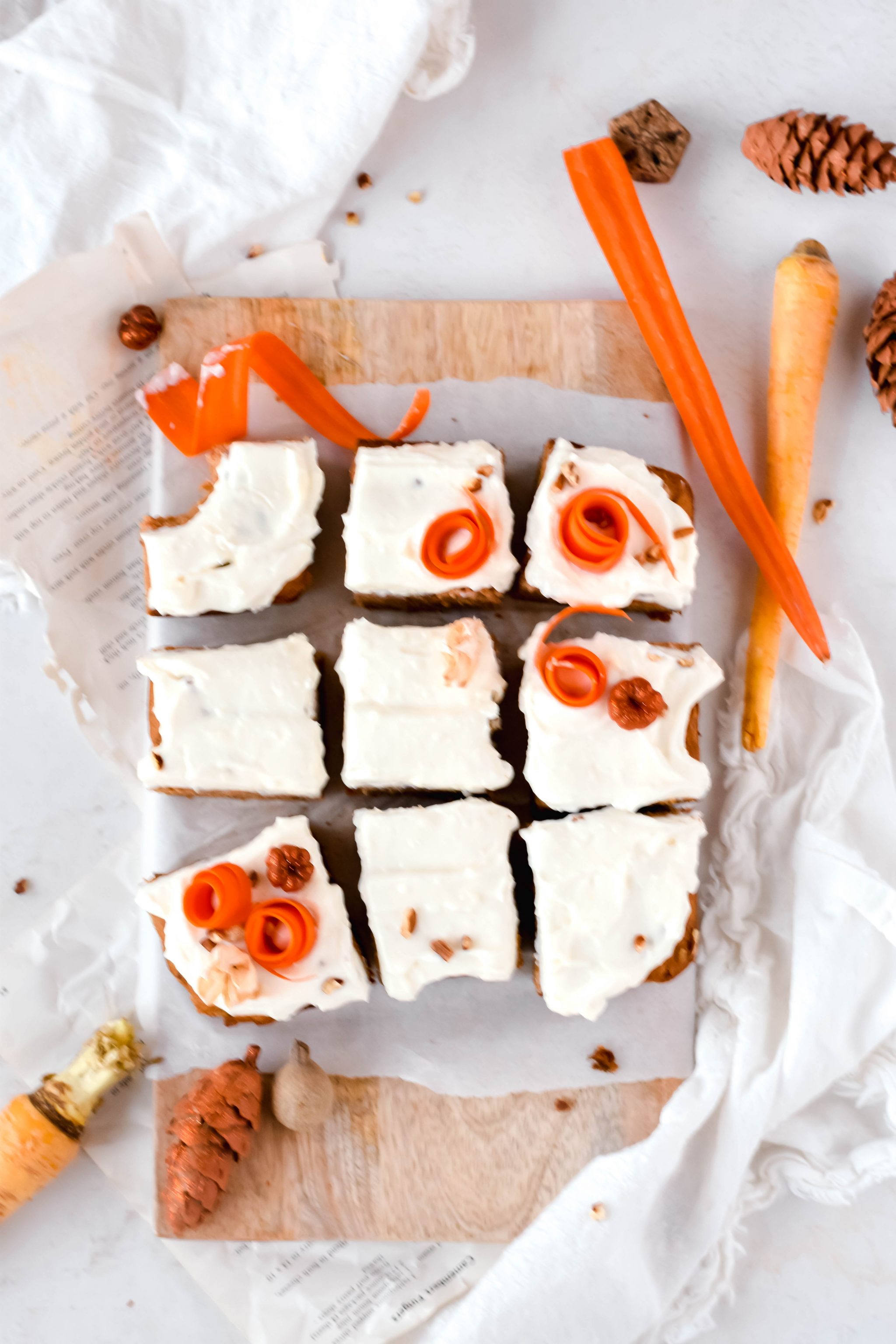 Overhead view of 9 square carrot cake bars topped with cream cheese icing and shredded carrots on a cutting board lined with parchment paper