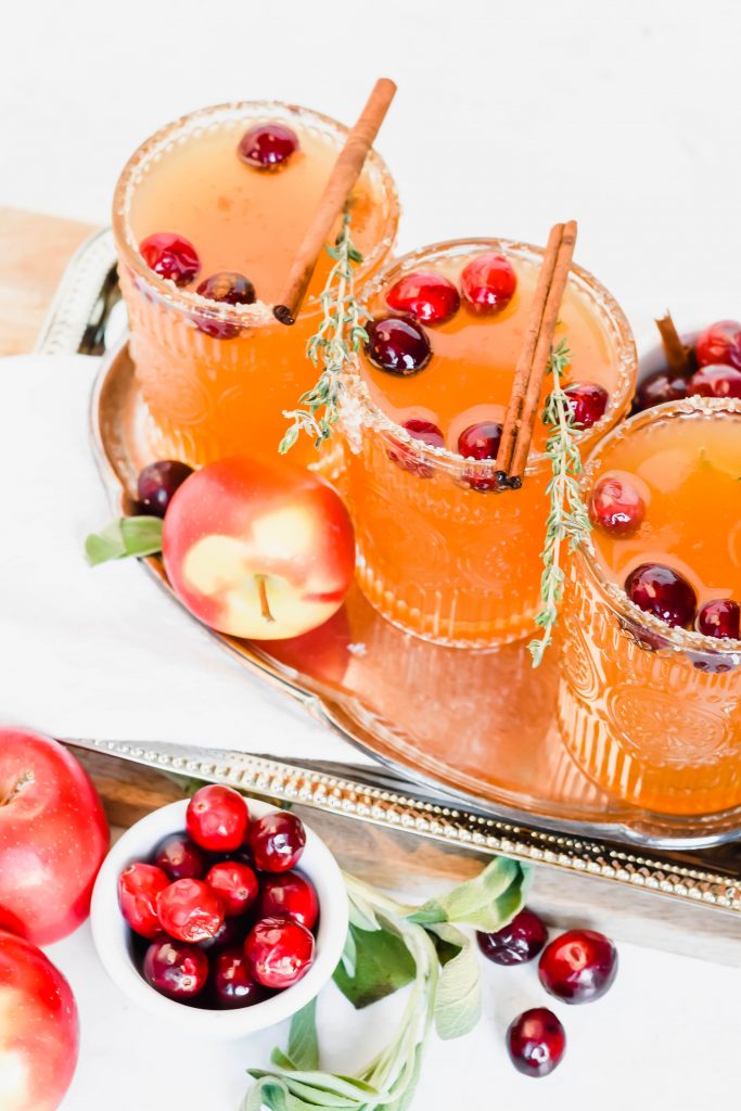 three garnished thanksgiving margaritas on silver tray surrounded by fresh herbs, cranberries, apples, and cinnamon sticks on white background