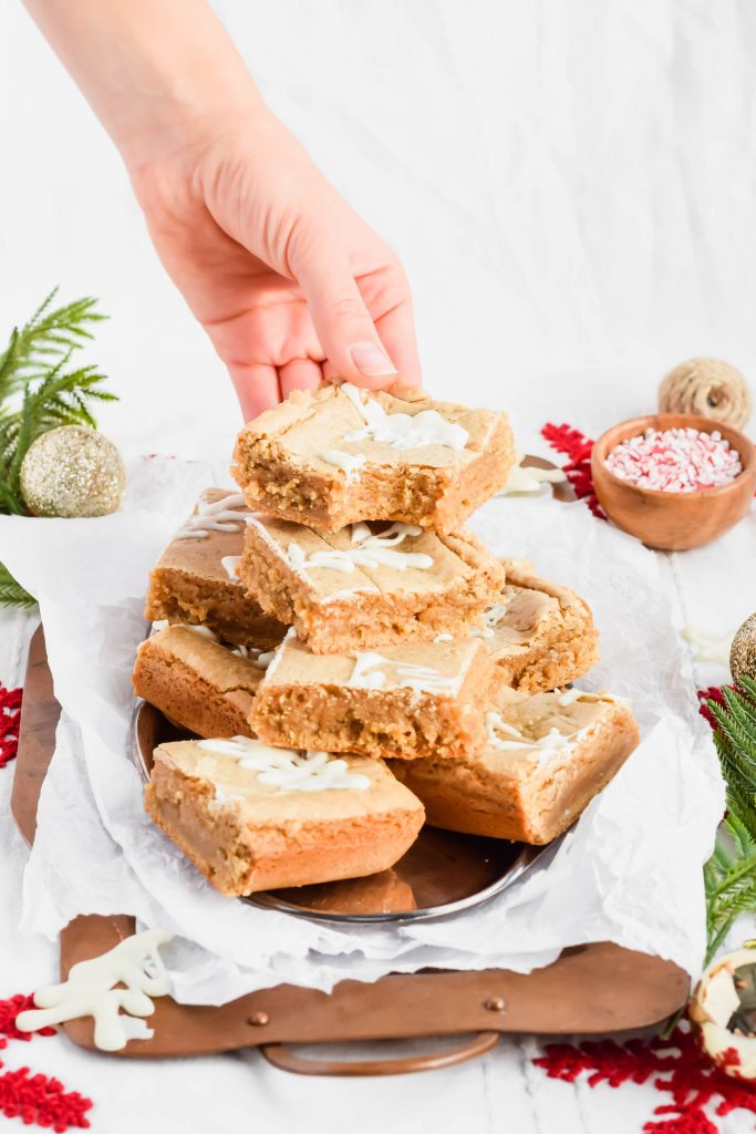sliced and stacked gingerbread blondies with hand lifting up the top blondie all stacked on silver tray on top of white parchment surrounded by Christmas decorations