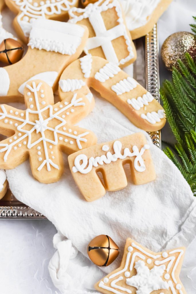 E shaped cookie shortbread christmas cookie with white icing writing emily on white linen sheet on top of a silver tray with additional cookies in background