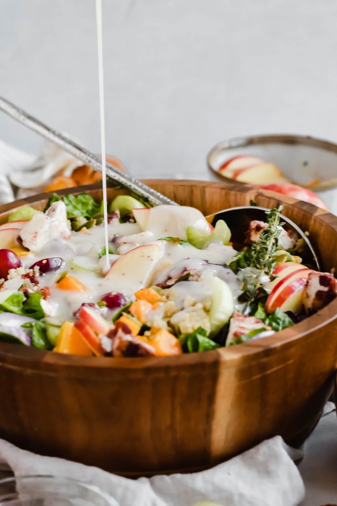 Pouring creamy white dressing on a Seasonal Harvest Salad in a wooden serving bowl with a silver salad spoon