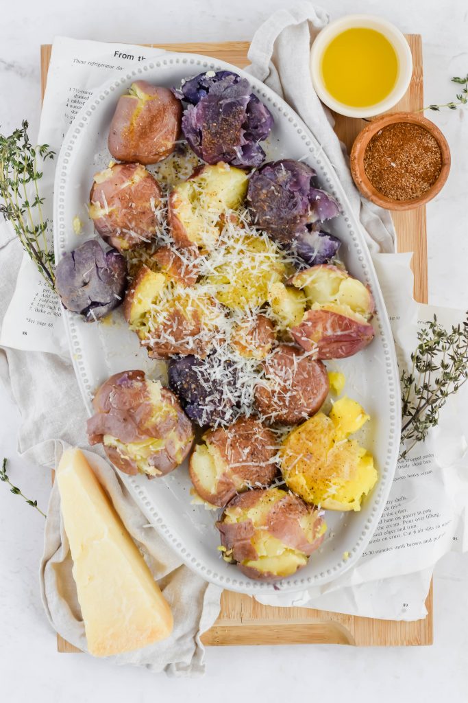 overhead shot of smashed potato bites on white plate with parmesan sprinkled on top surrounded by fresh herbs and additional recipe ingredients