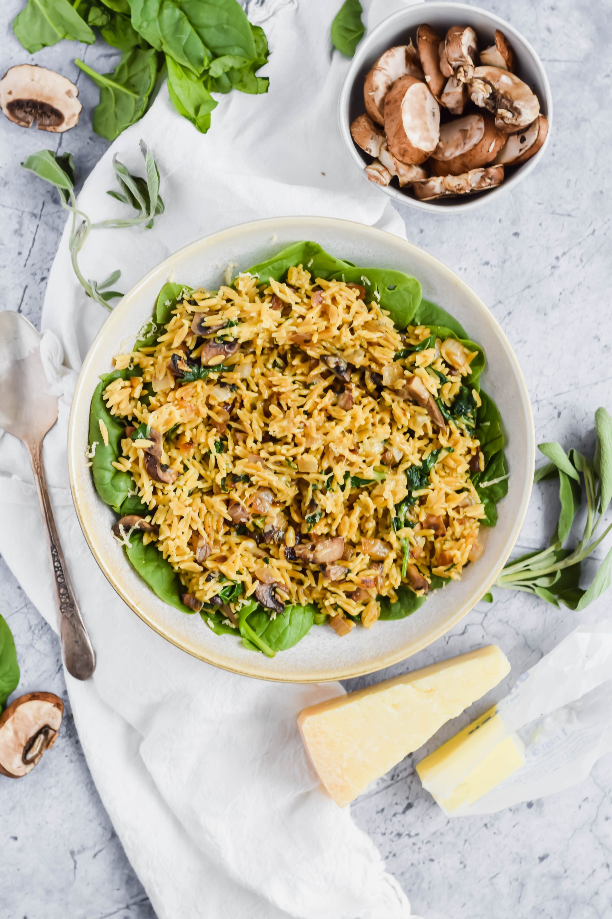 Large white bowl of Mushroom and Spinach Rice on a bed of spinach next to a silver serving spoon, fresh sage, butter, and a wedge of gouda cheese