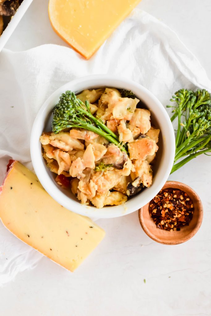 individual small white bowl of gouda mac and cheese next to block of gouda and small bowl of red pepper flakes and broccolini with white background