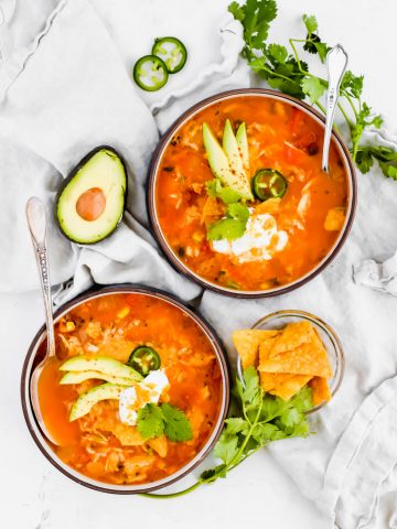Two bowls of Fiesta Chicken Tortilla Soup with spoons on white linen surrounded by avocado, jalapeno, cilantro, and chips