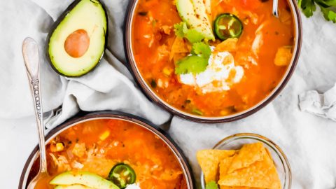 Two bowls of Fiesta Chicken Tortilla Soup with spoons on white linen surrounded by avocado, jalapeno, cilantro, and chips
