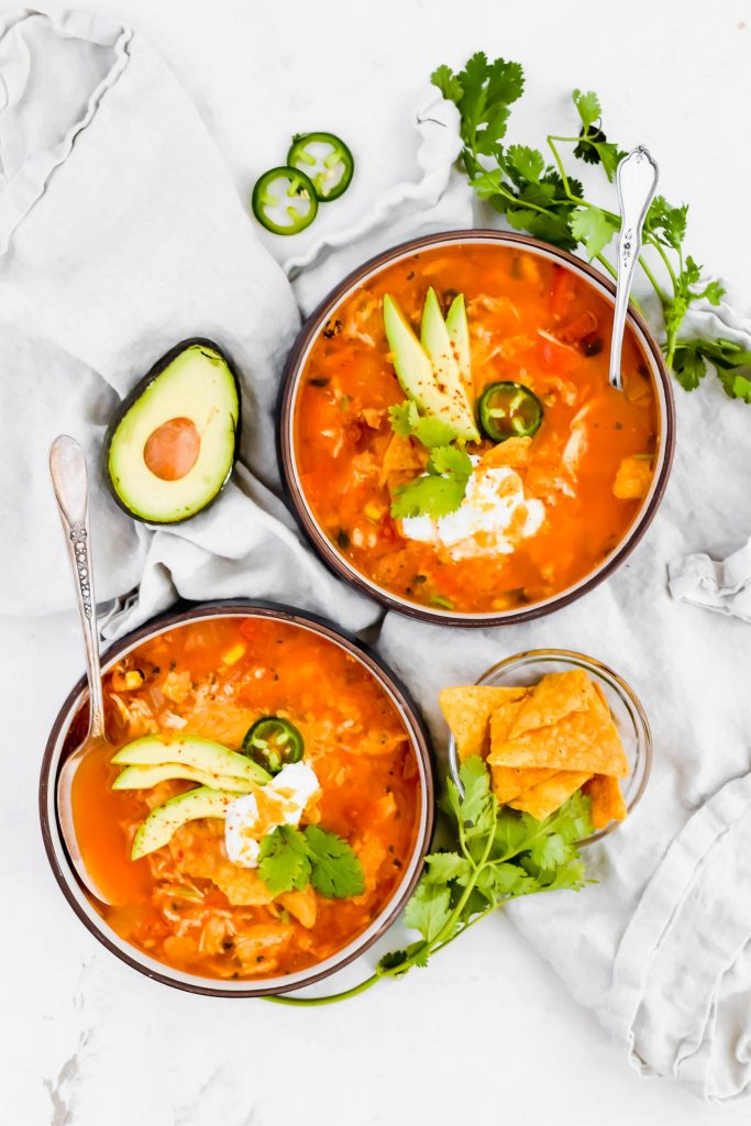 overhead shot of two bowls of fiesta chicken tortilla soup garnished and surrounded with avocado slices, cilantro, and jalapeno on white linen 