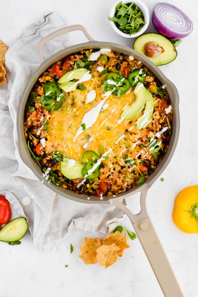 Overhead view of Cheesy Mexican Chicken Skillet in a tan pan surrounded by avocado, red onion, cilantro, chips, and bell pepper against a white linen backdrop