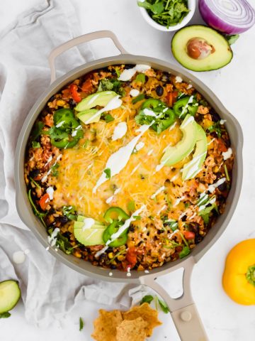 Overhead view of Cheesy Mexican Chicken Skillet in a tan pan surrounded by avocado, red onion, cilantro, chips, and bell pepper against a white linen backdrop