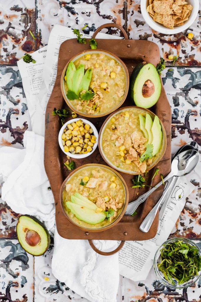 Three bowls of Dutch Oven White Chicken Chili recipe on a metal serving tray surrounded by small bowls tortilla chips, cilantro, and corn and avocado halves