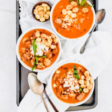 Three bowls of Spicy Sausage and Cauliflower Gnocchi Soup on white linen and a metal baking sheet with spoons and a small bowl of oyster crackers