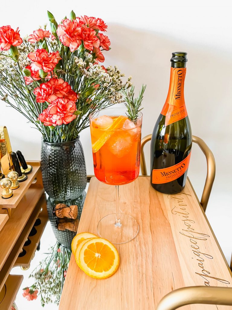 A rosemary aperol spritz cocktail on a wooden cutting board next to a bottle of prosecco and a bouquet of flowers