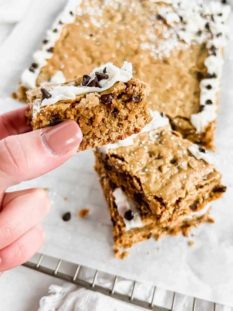 Holding an Oatmeal Chocolate Chip Blondie with frosting and chocolate chips on top in front of a blurred background of more blondies