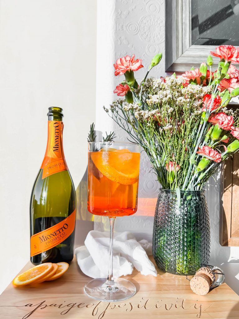 A rosemary aperol spritz cocktail on a wooden cutting board next to a bottle of prosecco and a bouquet of flowers