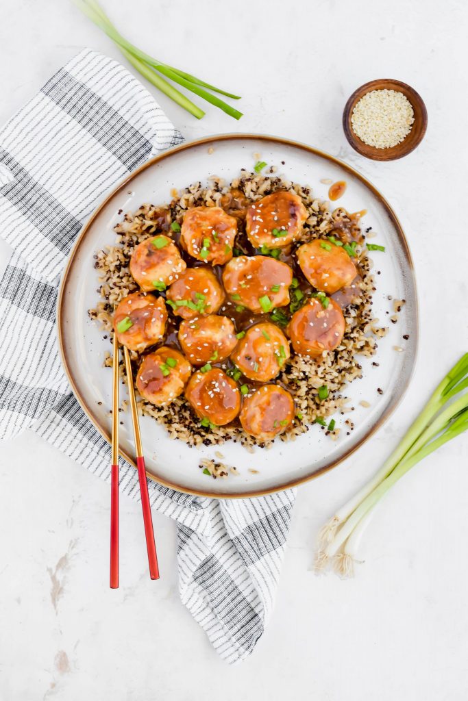 overhead shot of P.F. Chang's Cocktail Meatballs on white plate beside a small bowl of sesame seeds surrounded by additional green onion stalks with white background
