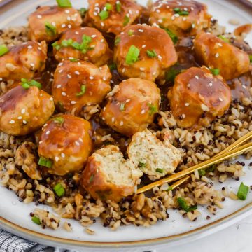 cocktail meatballs over bed of rice on white plate with chopsticks garnished with sesame seeds and herbs.