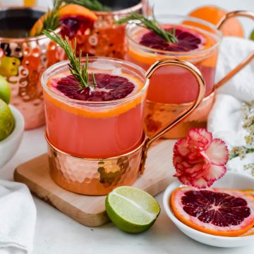Two Blood Orange Moscow Mules garnished with blood orange slices and fresh rosemary on a wooden cutting board surrounded by slices of blood orange and lime