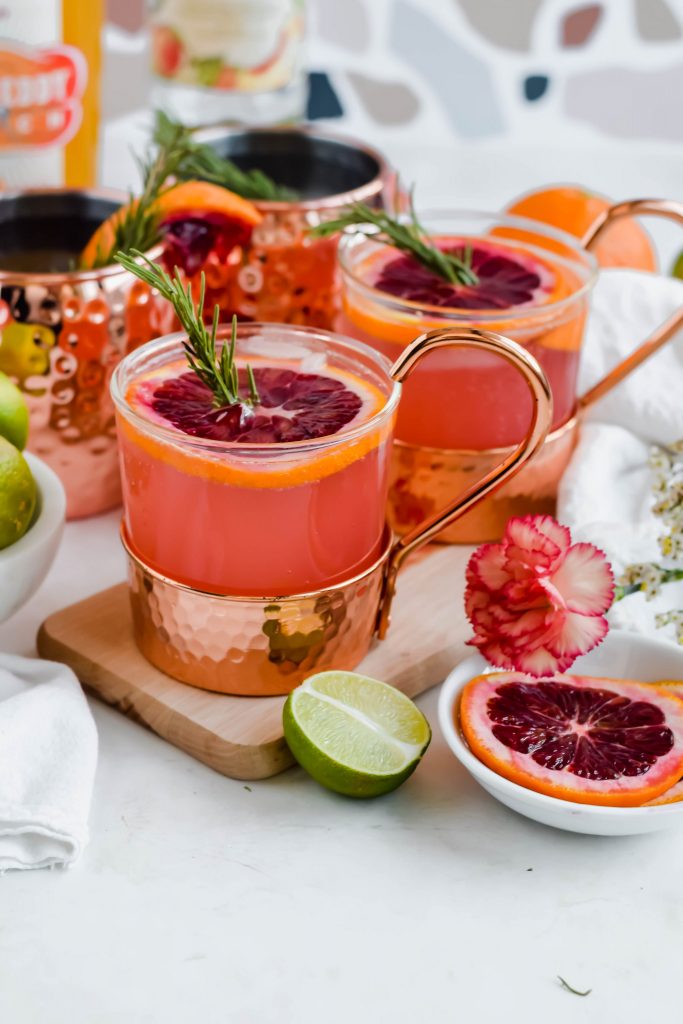 Two Blood Orange Moscow Mules garnished with blood orange slices and fresh rosemary on a wooden cutting board surrounded by slices of blood orange and lime