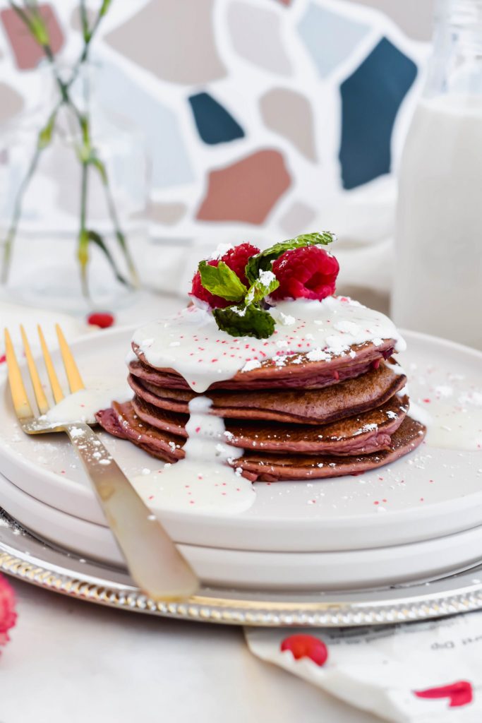 Stack of Healthy Red Velvet Protein Pancakes on a white plate with a gold fork topped with cream cheese frosting and fresh berries