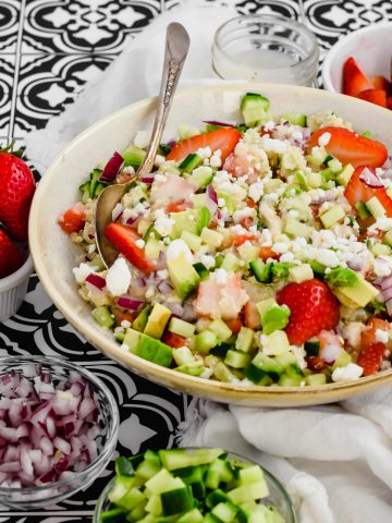 Overhead view of a big salad bowl filled with Strawberry Quinoa Salad surrounded by a bowl of red onion, a bowl of feta cheese, a bowl of strawberries, a bowl of cucumbers, and a small container of dressing