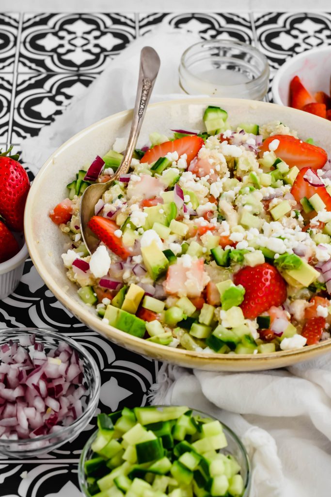 Overhead view of a big salad bowl filled with Strawberry Quinoa Salad surrounded by a bowl of red onion, a bowl of feta cheese, a bowl of strawberries, a bowl of cucumbers, and a small container of dressing