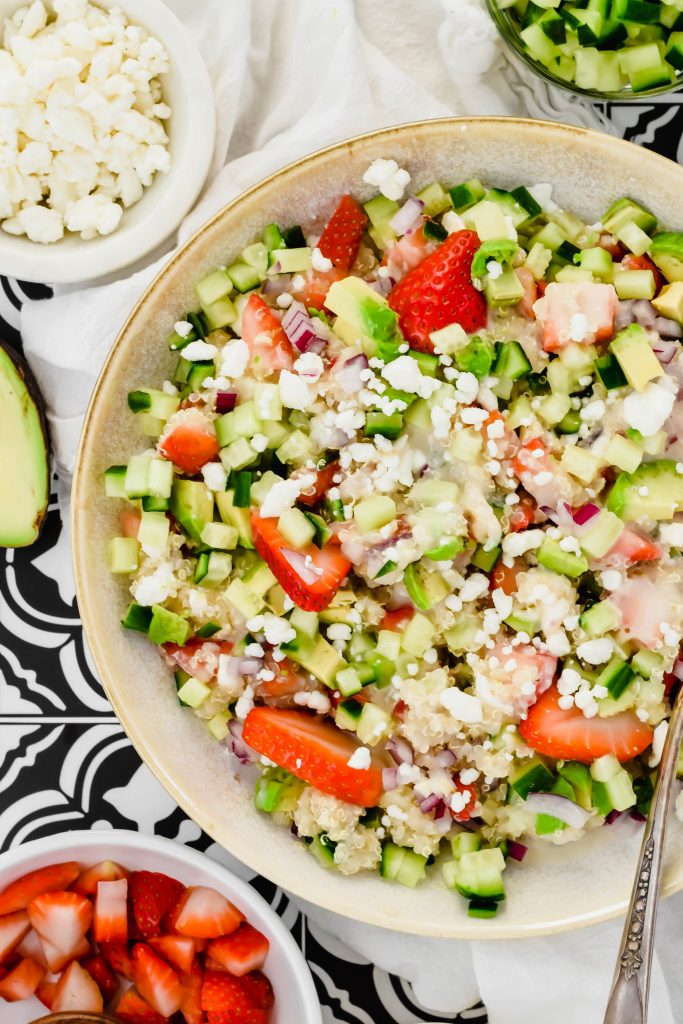 Overhead view of a big salad bowl filled with Strawberry Quinoa Salad surrounded by half an avocado, a bowl of feta cheese, a bowl of strawberries, a bowl of cucumbers, and a small container of dressing